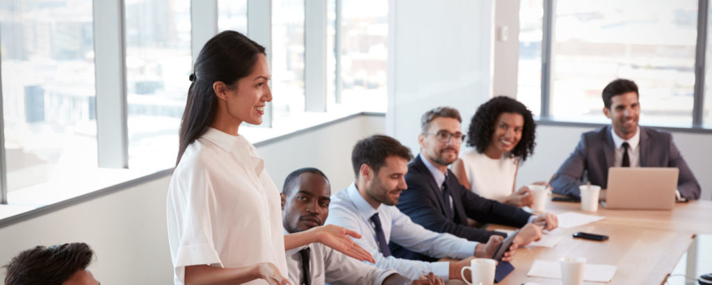 Businesswoman Stands To Address Meeting Around Board Table