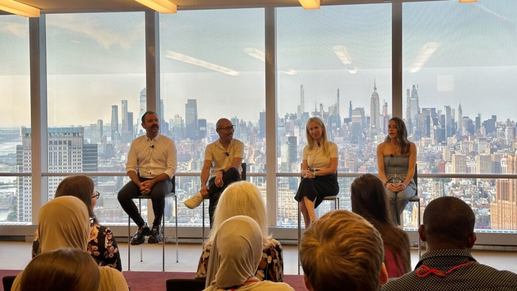 Four people sitting on stools talking to a crowd of people with large glass windows behind speakers displaying NYC skyline in the distance.