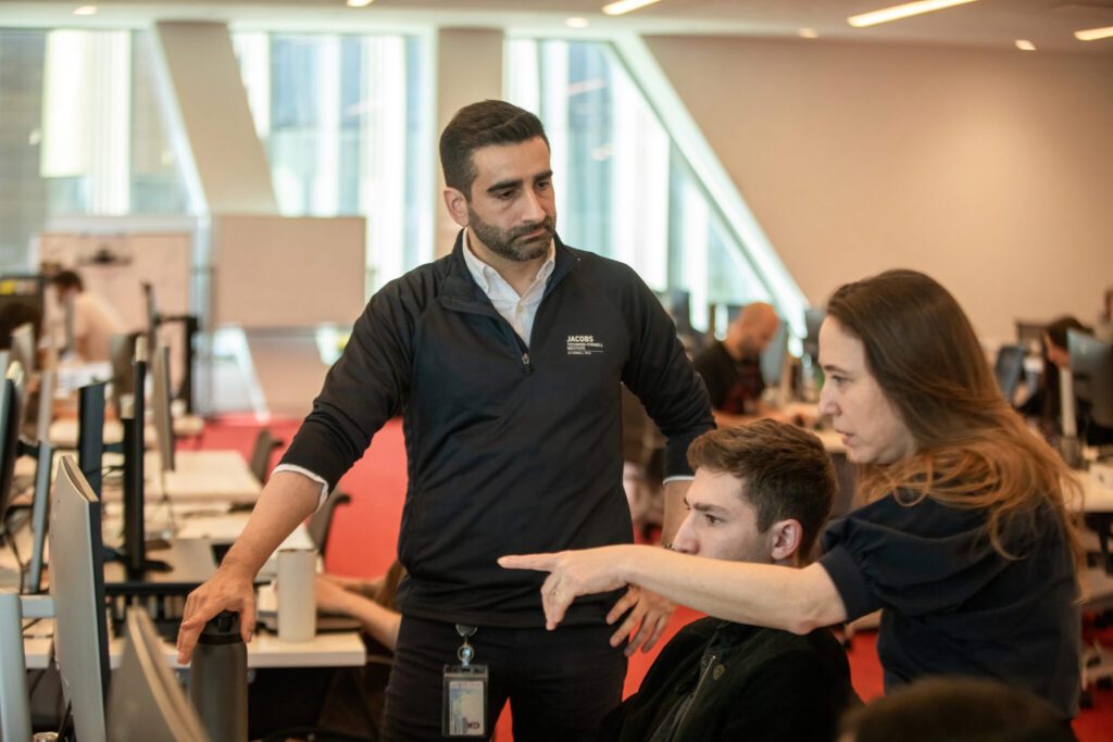 man with dark hair wearing a dark 3/4 zip sweater talking to other people pointing at a computer screen.