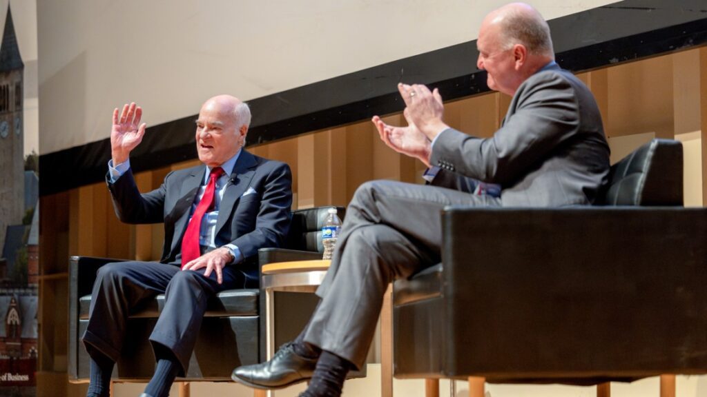 Two men wearing business suits sitting in chairs on a stage, one clapping and the other waving at the audience.