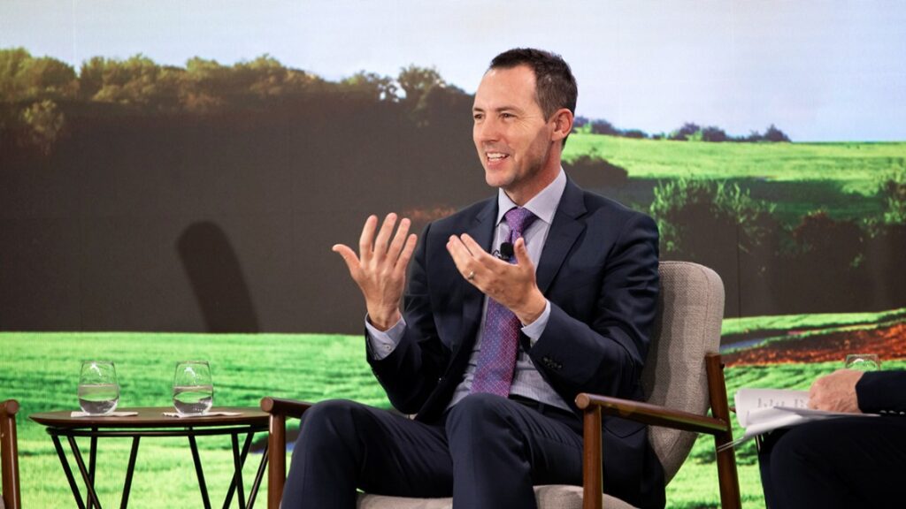 Man in black suit sitting in chair with field of green background, hands up and smiling.