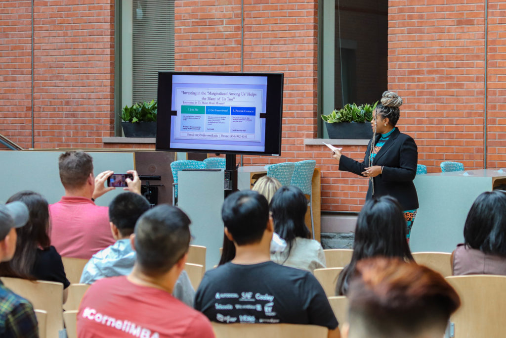 A woman pitches her business in front of a screen to an audience in an atrium space