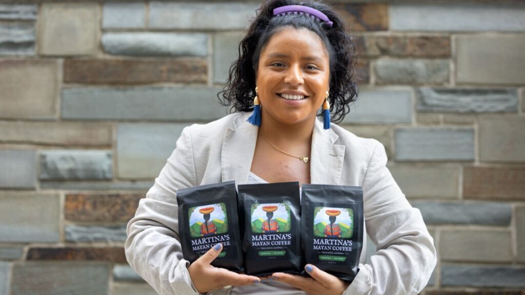 Woman with dark hair pulled up with a purple headband wearing a beige sport coat and teal dangly earrings with her arms full of packaged coffee beans.