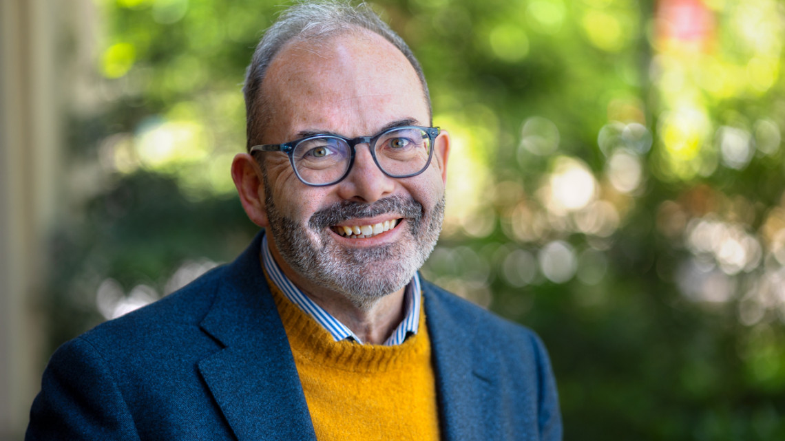 man with glasses and a yellow sweater under sport coat with a greenery background