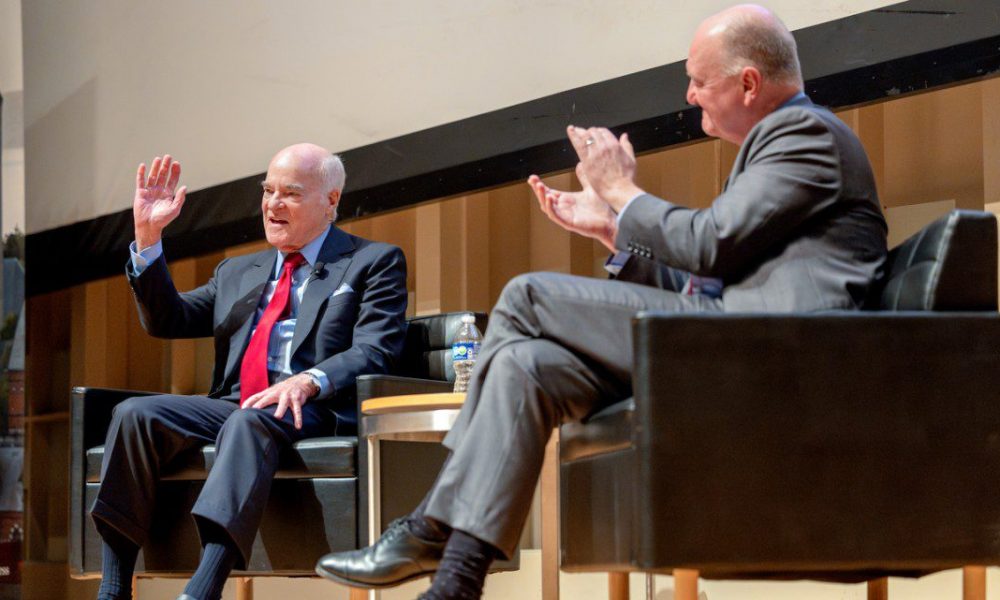 Two men wearing business suits sitting in chairs on a stage, one clapping and the other waving at the audience.