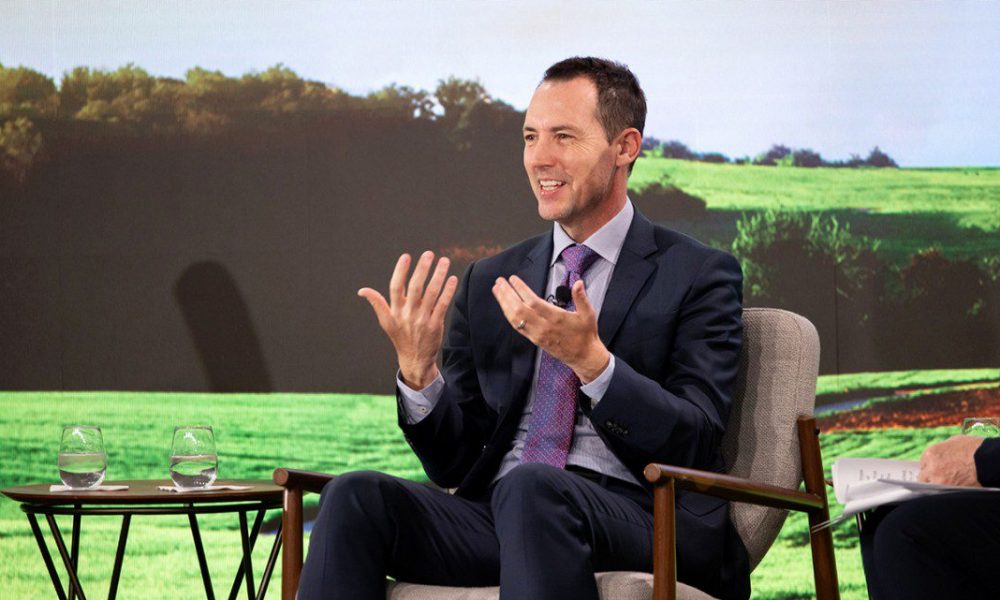 Man in black suit sitting in chair with field of green background, hands up and smiling.