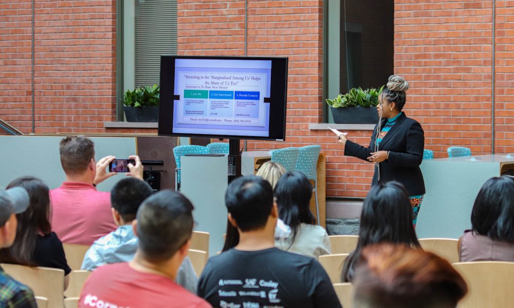 A woman pitches her business in front of a screen to an audience in an atrium space
