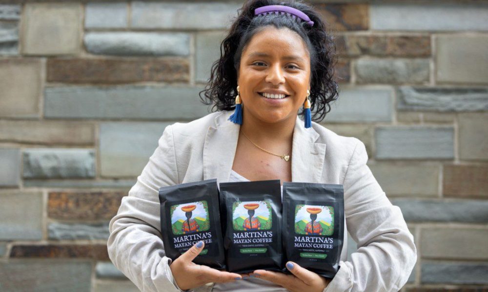 Woman with dark hair pulled up with a purple headband wearing a beige sport coat and teal dangly earrings with her arms full of packaged coffee beans.