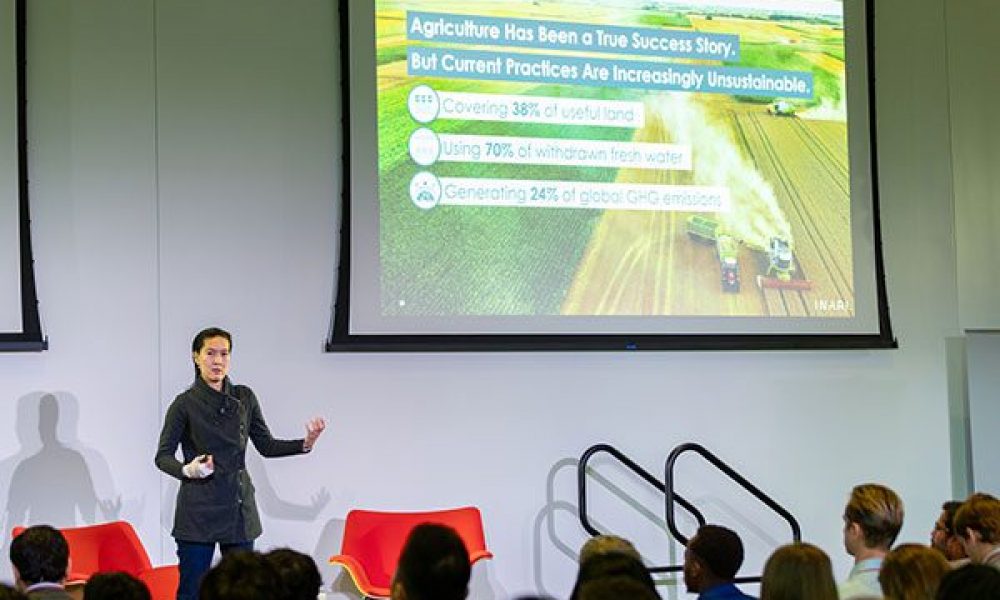 Person talking in front of audience with presentation screen in background.