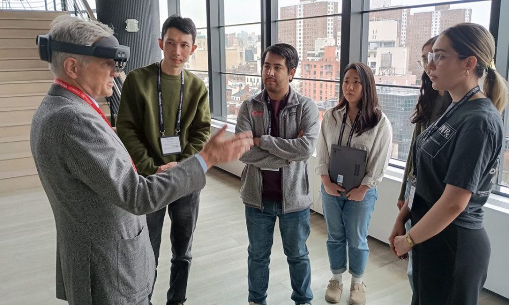 Group of people standing together looking at a man in a suit wearing headgear.