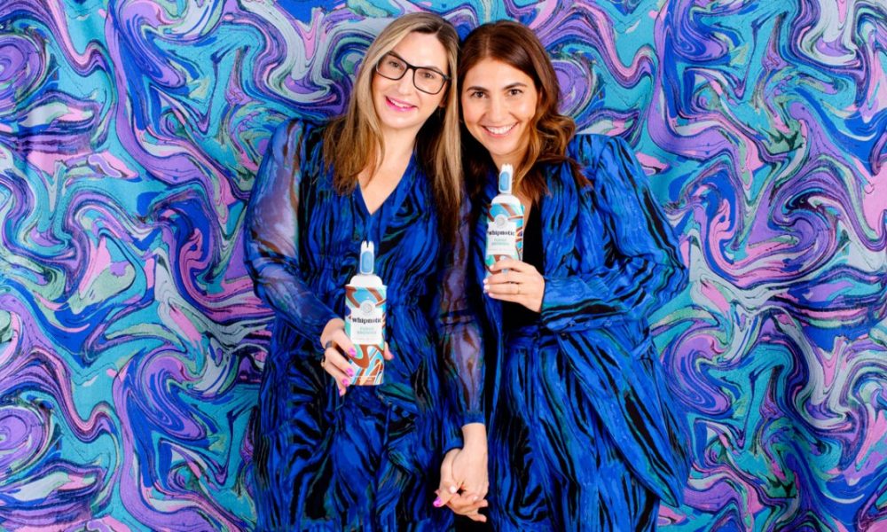 Two woman, both wearing blue dresses, one with blonde hair, one with brunette hair, holding cans of whipped cream standing in front of a blue, purple, turquoise, and lavender tie dyed background