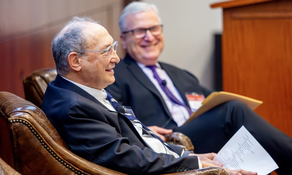 two older men wearing suits sitting in brown leather chairs having a discussion, smiling
