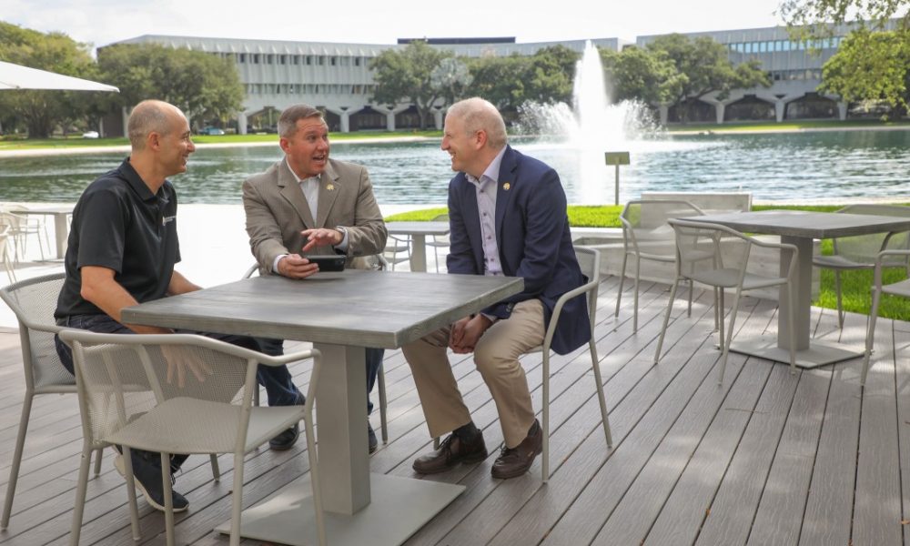 Three men sitting outside at patio table having a discussion, water fountain and buildings in the background.