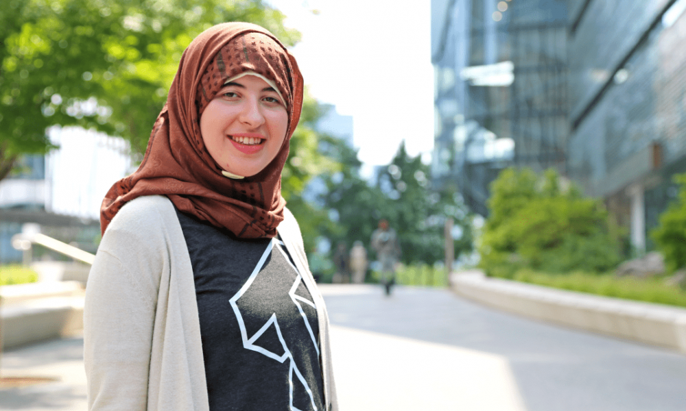 Woman wearing a Cornell Tech t-shirt, cardigan, and head scarf with Cornell Tech in background.