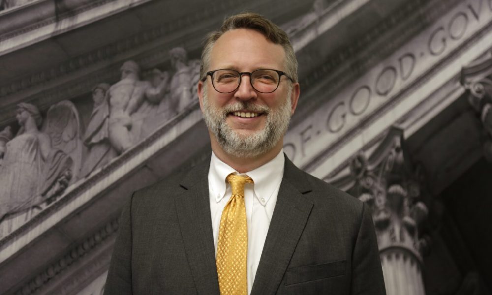 Person wearing a dark suit with white shirt and yellow tie and glasses, with blurred background of ancient architecture