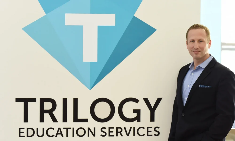 Dan Sommer stands in front of a wall with the logo of Trilogy Education Services