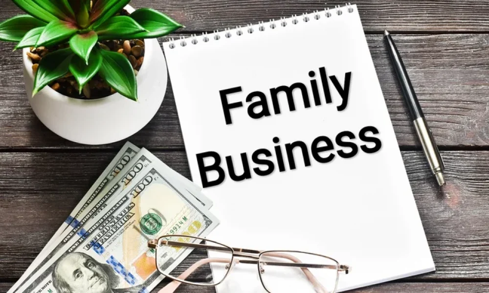 The words Family Business on the front of a top spiral notebook with one hundred dollar bills, eyeglasses, a pen, and a plant next to it.