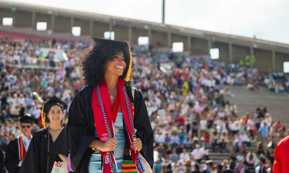 College graduate wearing multiple honor tassels, cap and gown, with large crowd blurred in the background