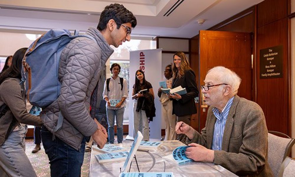 man in sport jacket and blue shirt wearing glasses signing books for line of students.