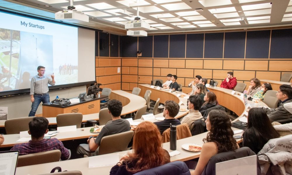 Lecture style classroom filled with students watching speaker in front of room standing in front of large screen giving presentation