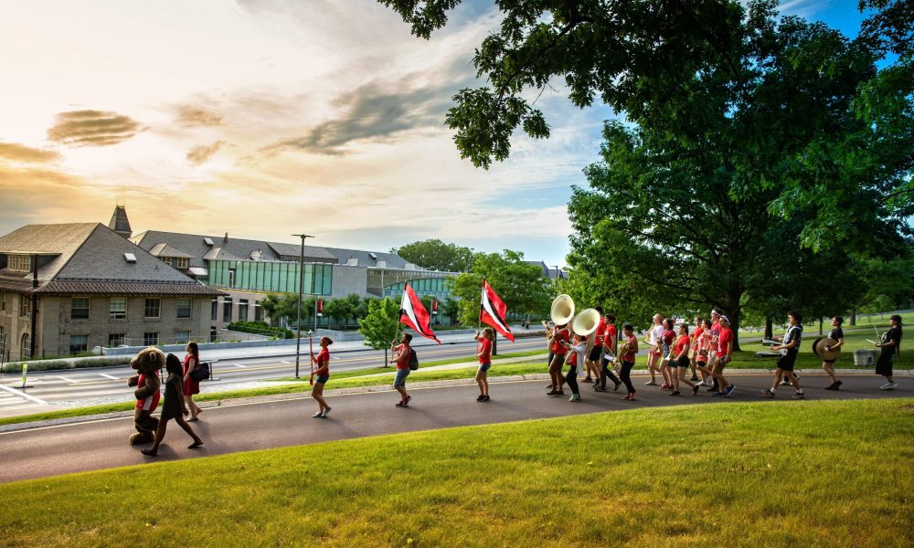 Cornell University marching band led by University mascot, Bear in jersey. All members walking down slope with campus scene in background.