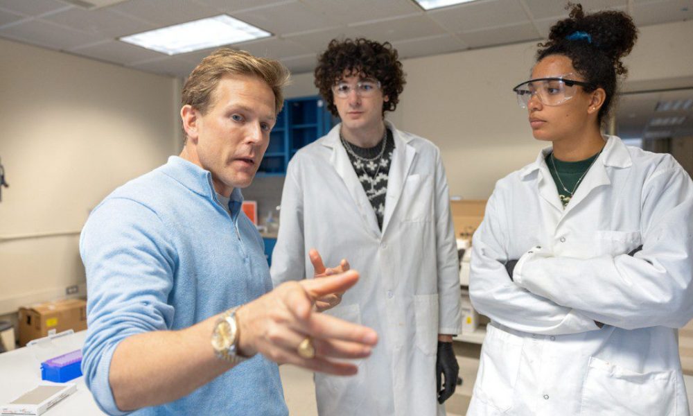 three people in a lab, one in a blue shirt pointing closely at the camera