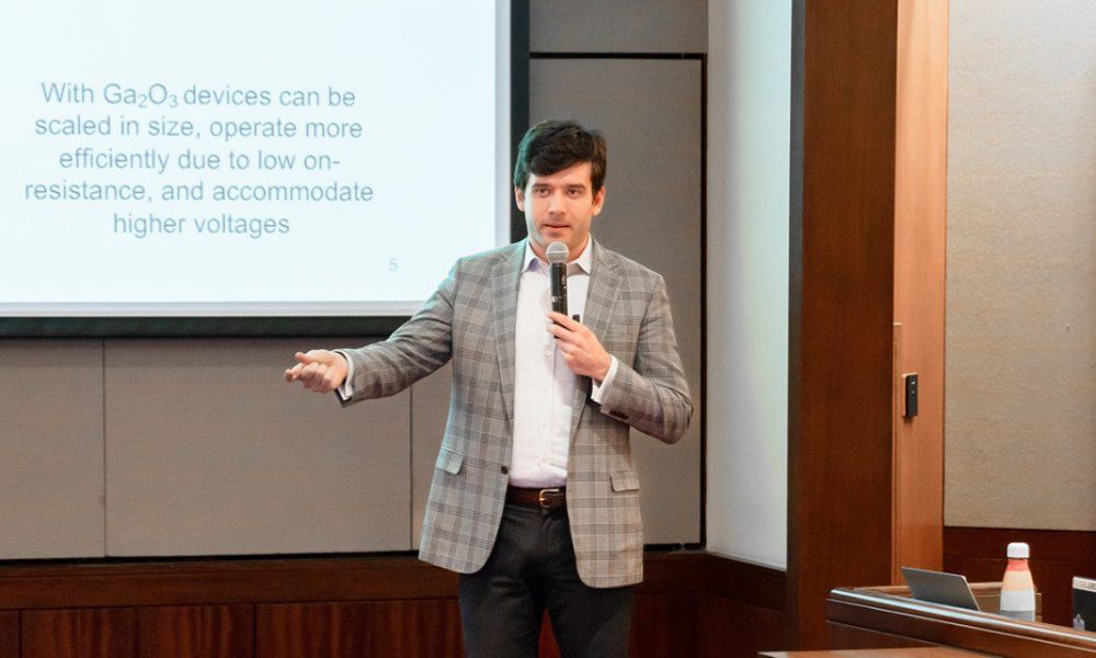 man standing in front of presentation screen, wearing plaid blazer, speaking into a microphone with right hand and arm raised out.