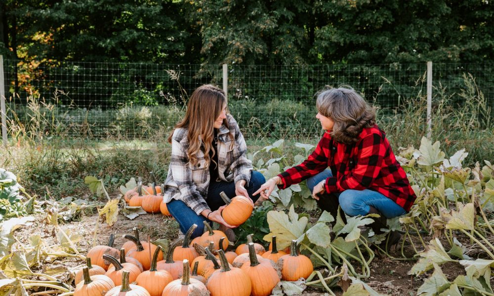 Two woman wearing plaid flannel shirts kneeling in a pumpkin patch