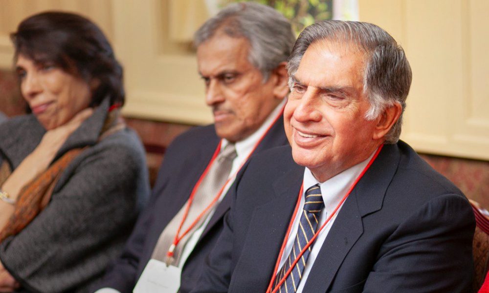 Ratan Tata wearing suit and tie sitting and smiling, side angle. People sitting next to him blurred in the background.