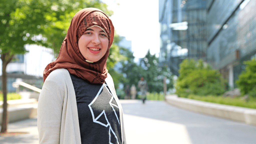 Woman wearing a Cornell Tech t-shirt, cardigan, and head scarf with Cornell Tech in background.