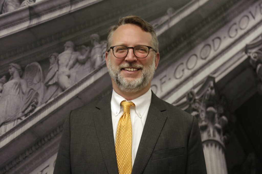 Person wearing a dark suit with white shirt and yellow tie and glasses, with blurred background of ancient architecture