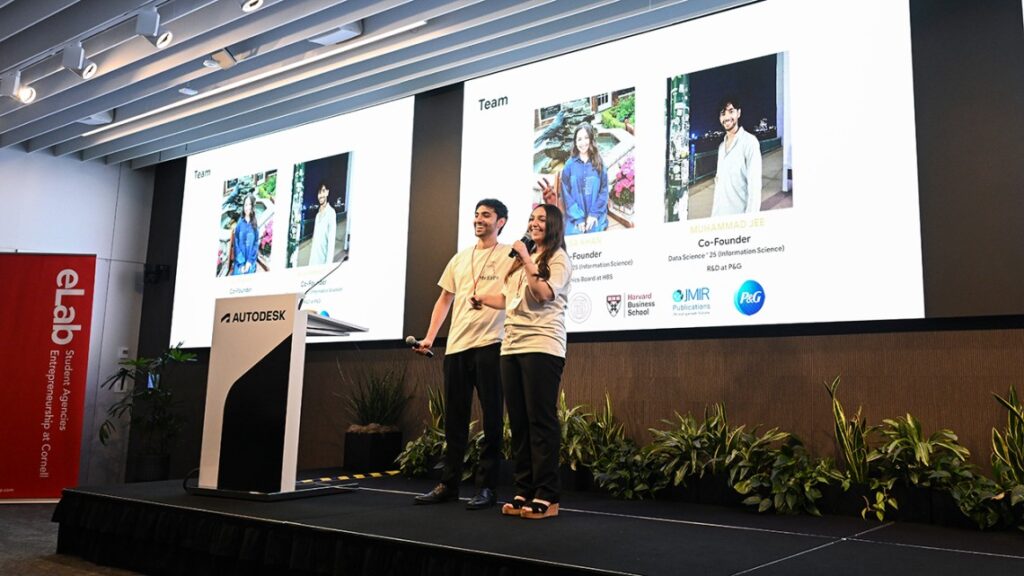 Two people standing on a stage wearing beige t-shirts with presentation screens behind them