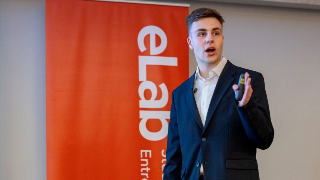 Man in business suit holding remote standing in front of a red vertical banner