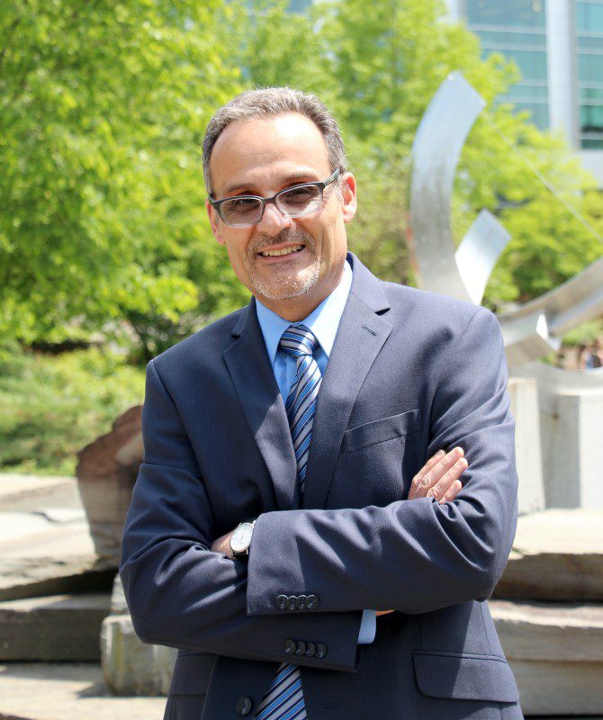 man wearing suit and tie standing with his arms crossed with greenery in the background