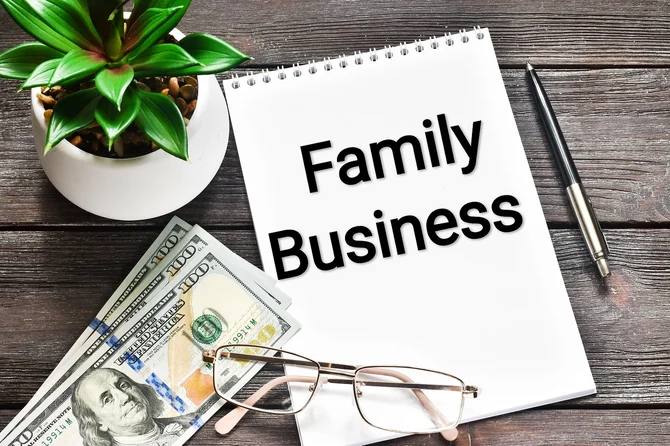 The words Family Business on the front of a top spiral notebook with one hundred dollar bills, eyeglasses, a pen, and a plant next to it.