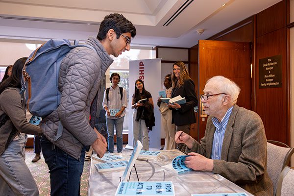 man in sport jacket and blue shirt wearing glasses signing books for line of students.