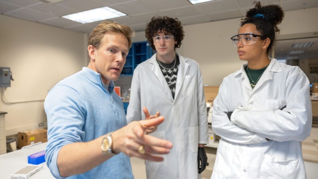 three people in a lab, one in a blue shirt pointing closely at the camera