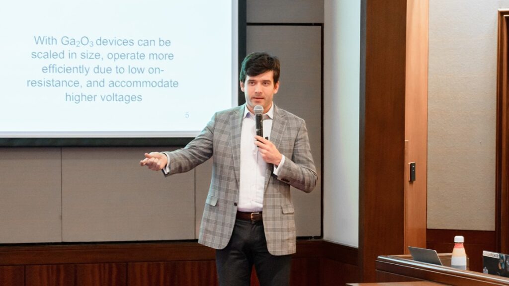 man standing in front of presentation screen, wearing plaid blazer, speaking into a microphone with right hand and arm raised out.