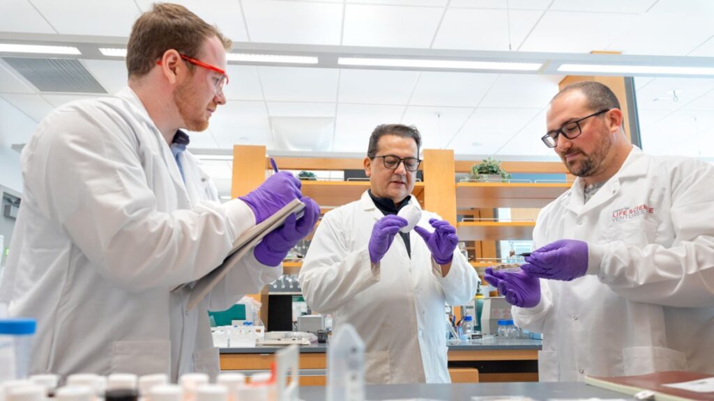 Three men, all wearing glasses in standing in a science lab, wearing white lab coats and purple safety gloves.