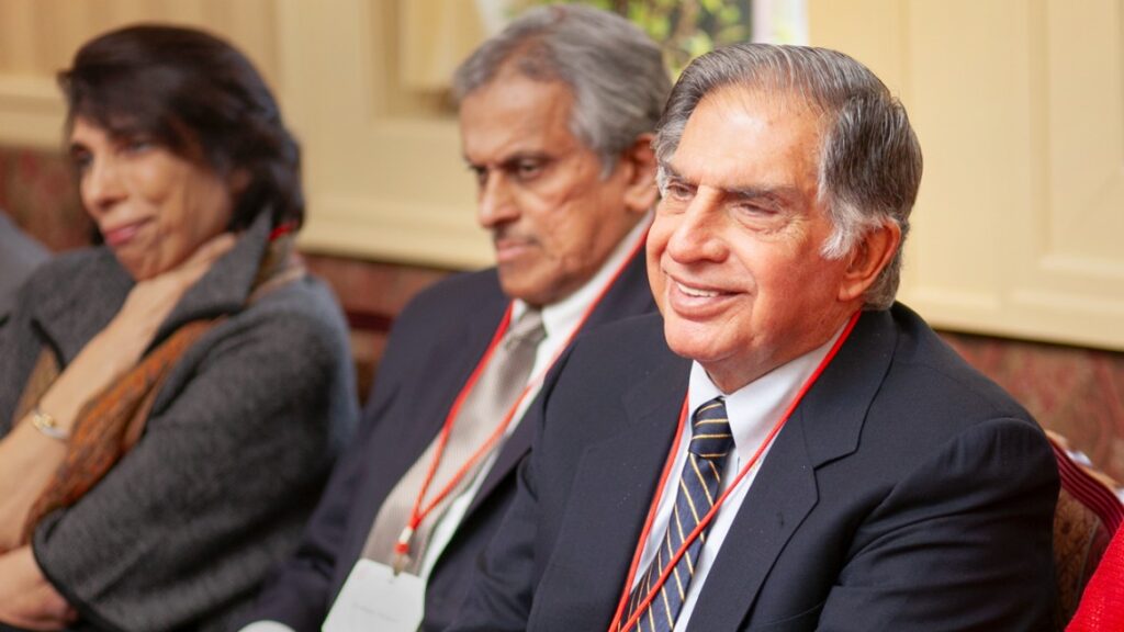 Ratan Tata wearing suit and tie sitting and smiling, side angle. People sitting next to him blurred in the background.